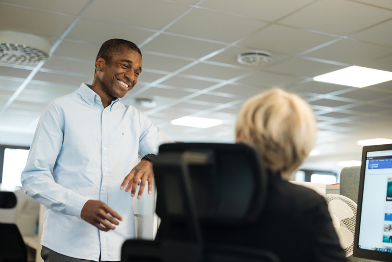 Två medarbetare samtalar med varandra, en ung kille står upp vid sidan av ett skrivbord där en kvinna sitter ner vid sin dator så man ser hennes ryggtavla bara. 