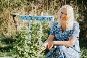 En kvinnas i blå klänning sitter bland grönska och ler. Bakom henne syns en skylt där det står Safirträdgården. 