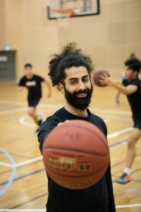 En man håller en basketboll mot kameran. I bakgrunden spelar två killar basket.
