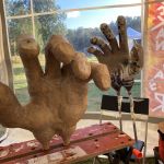 I ett tält sitter två jättestora dockhänder fast i arbetsbänkar. Bredvid ligger en sommarhatt.