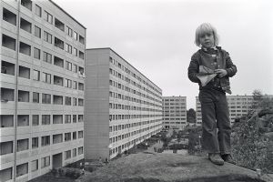 En svartvit bild på en pojke på ett berg med miljonprogramhus i bakgrunden.