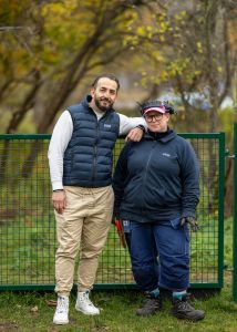 En man och en kvinna i arbetskläder ler mot kameran. I bakgrunden en hundrastgård.