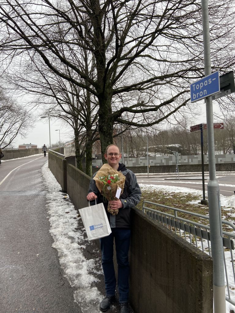 Hyresgäst står vid Topasbron och håller i blommor och diplom. 