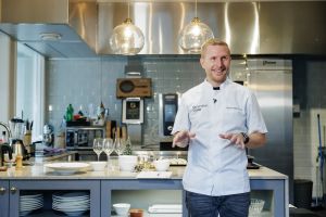 En mat i kockkläder med ett kök i bakgrunden.