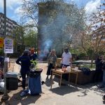 Grillaktivitet i en park. I bakgrunden synt träd och höghus i Landala. Det ryker från grillen och det står ketchup på bordet.