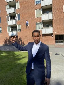 En stilig ung man i kavaj och kort lockigt svart hår ser glad ut och pekar mot en av karaktärerna som ingår i utställningen x-times people chair och hänger på fasaden bakom honom.