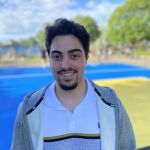 En glad ung man med mustasch och skägg klädd i grå luvtröja och randig pikétröja8 står framför basketplanen vis Svarte Mosse.