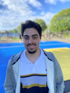 En glad ung man med mustasch och skägg klädd i grå luvtröja och randig pikétröja8 står framför basketplanen vis Svarte Mosse. 