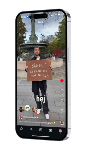 En mobiltelefon och på bildskärmen ser man en kille som håller en skylt där det står Säg hej! Få kaffe och kanelbulle.