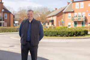 En man ler in i kameran med röda hus i bakgrunden.