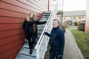 En kvinna i en trappa och en man i arbetskläder bredvid tittar in i kameran.