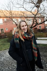 En kvinna ler in i kameran. I bakgrunden hus och träd.