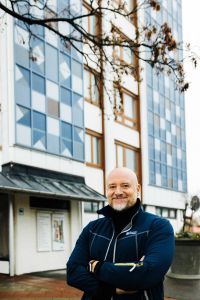 En man står med armarna i kors och ler in i kameran med en husfasad i bakgrunden.