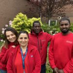 Gruppbild på fyra trygghetsvärdar i röda jackor. Två kvinnor och två män. De står framför blommande buskar.