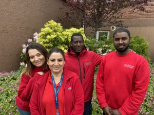 Gruppbild på fyra trygghetsvärdar i röda jackor. Två kvinnor och två män. De står framför blommande buskar.