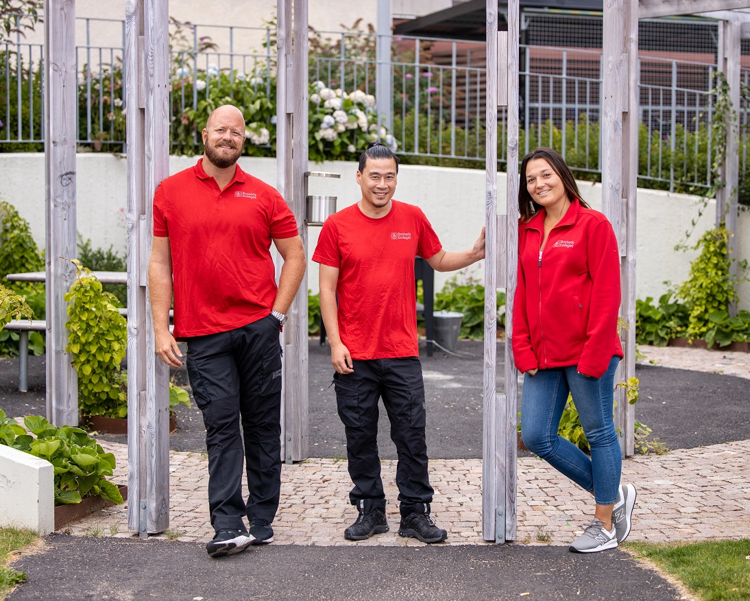 Tre trygghetsvärdar i röda tröjor står bredvid varandra och ler in i kameran.
