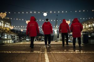 Man ser ryggen på fyra Trygghetsvärdar i röda vinterjackor. De går bort från bild.