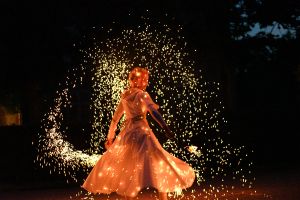 En kvinna med ljust kort hår snurrar runt i ett regn av lysande gnistor i mörkret så att hennes vita klänning står ut.