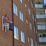 en karaktär i utställningen x-times people chair pratar i telefon och äter jordgubbar på en stol som är fastskruvad i fasaden. Två personer fotar honom från sina balkonger.