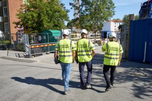 Tre män går ner mot en byggarbetsplats. De har gula västar på sig där det står Rättvist Byggande på ryggen.