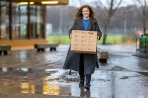 En kvinna med en flyttlåda i famnen går mot kameran och ler.