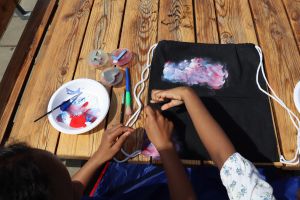 Tre händer gör ett tygtryck i blått, vitt och rosa på ett svart tyg mot ett träbord.