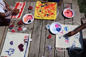 Händer som gör tygtryck i blått, rosa och rött på vit och gul bakgrund på ett träbord.