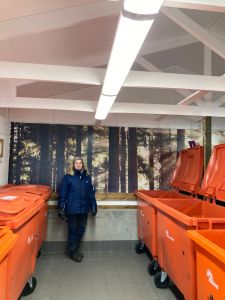 En kvinna står och tittar in i kameran i ett miljörum med sopkärl på sidorna och en fototapet i bakgrunden.