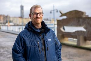 En man står utomhus och ler in i kameran i arbetskläder.