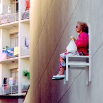 Äldre kvinna sitter på en vit stol högt upp på en husfasad och äter ett äpple. Hon har en röd jacka och solglasögon på sig.