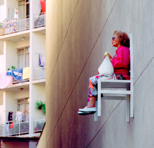 Äldre kvinna sitter på en vit stol högt upp på en husfasad och äter ett äpple. Hon har en röd jacka och solglasögon på sig.